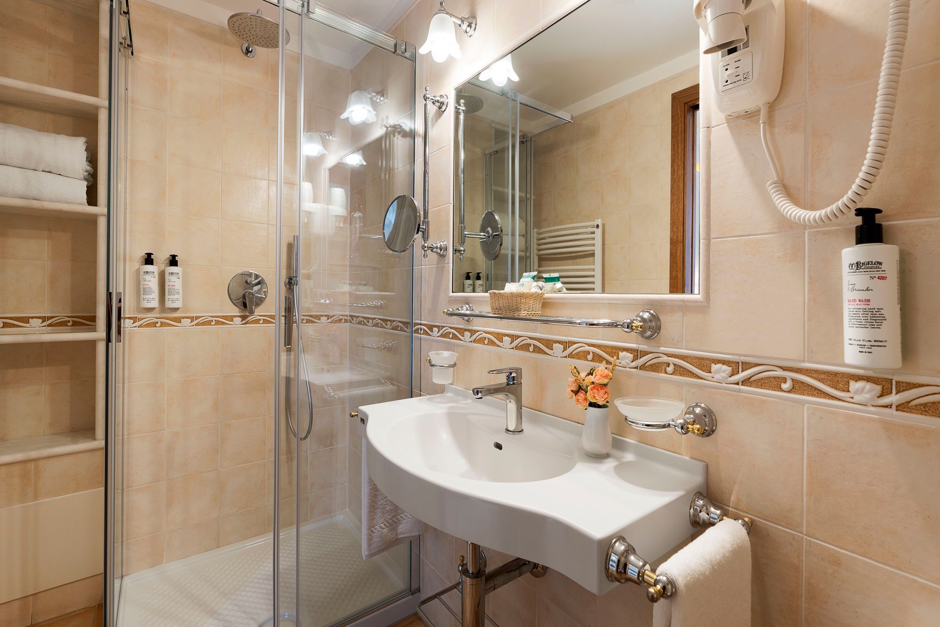 Kleines beigefarbenes Badezimmer mit Glasdusche, Waschbecken, Spiegel und wandmontierten Toilettenartikeln.