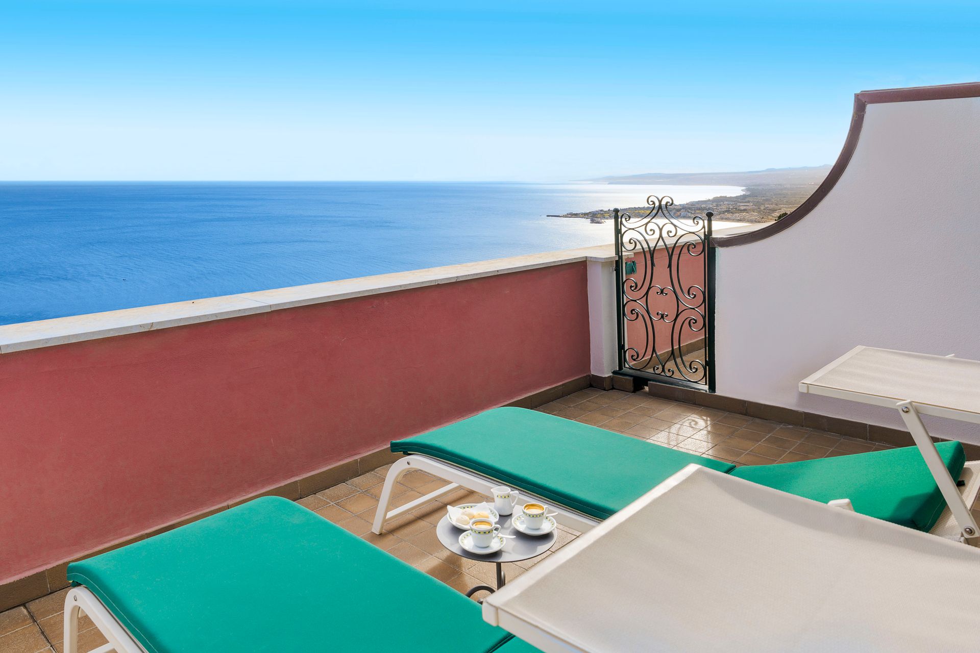 Balkon mit Meerblick, türkisfarbenen Liegestühlen, einem kleinen Tisch und roten Wänden unter blauem Himmel.