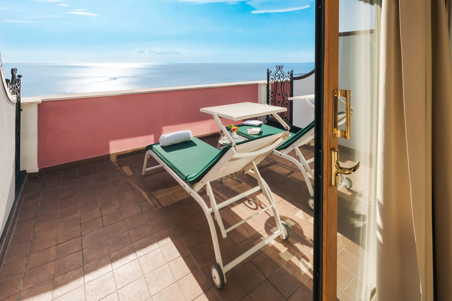Balkon mit zwei grünen Liegestühlen, einem Tisch und Meerblick durch eine offene Tür