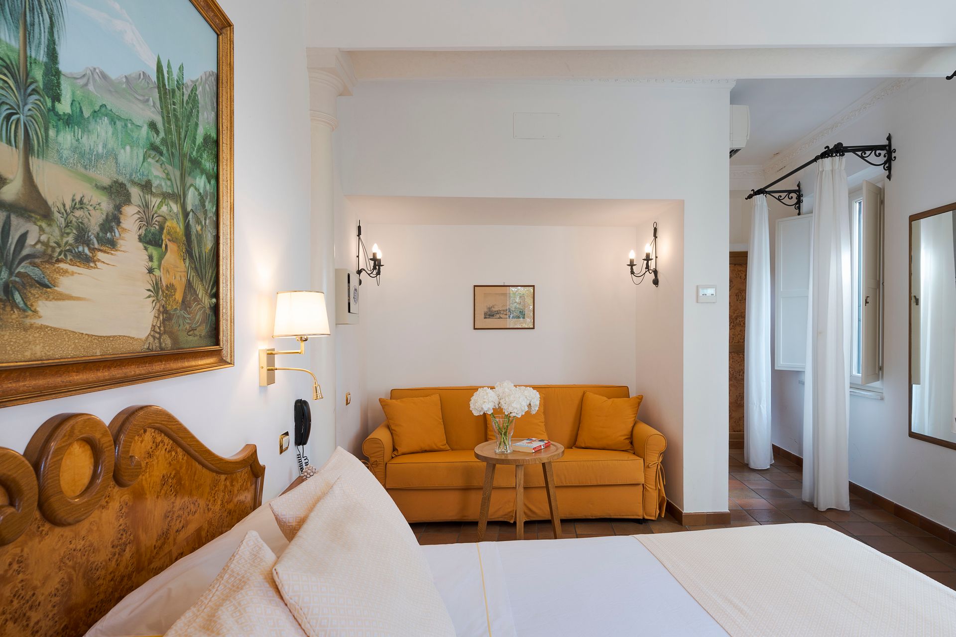 Schlafzimmer mit geschnitztem Holzkopfteil, weißer Bettwäsche, orangefarbenem Sofa und Wandbildern in einem hellen Zimmer