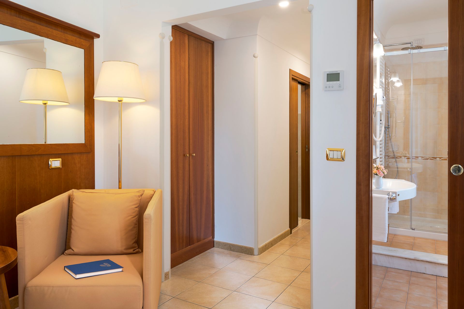 Hotelflur mit beigem Sessel, Lampe, Spiegel und Badezimmertür mit sichtbarer Dusche