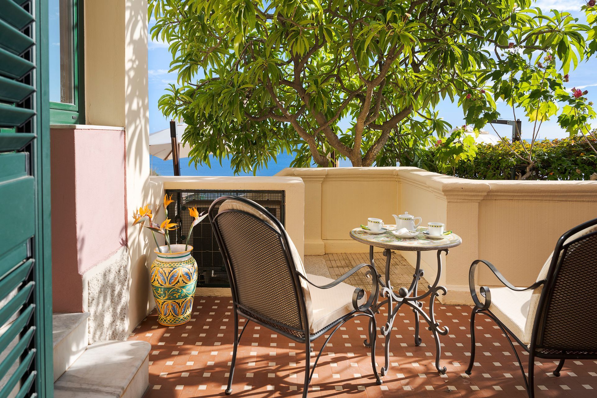 Sonniger Balkon mit Korbstühlen, kleinem Tisch und Topfpflanzen neben einer Hausfassade.
