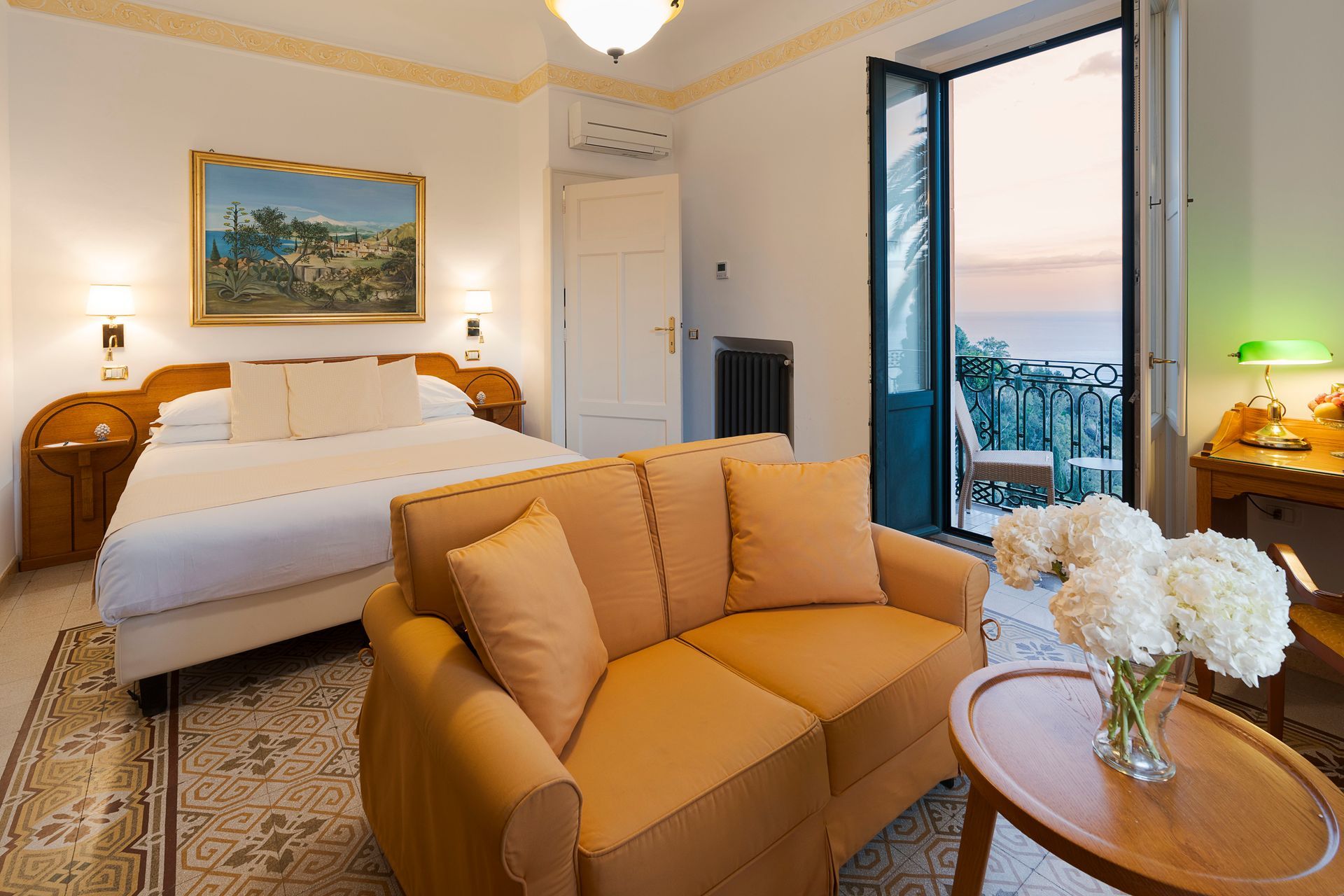 Helles Hotelzimmer mit Bett, beigem Sofa, rundem Tisch und offenen Balkontüren mit Meerblick.