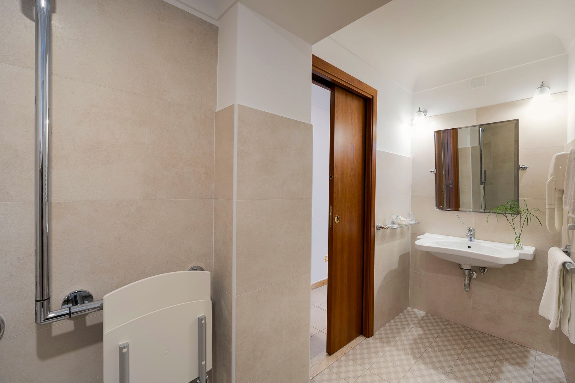 Kleines Badezimmer mit beigefarbenen Fliesen, wandhängender Toilette, Waschbecken, Spiegelschrank und Holztür.
