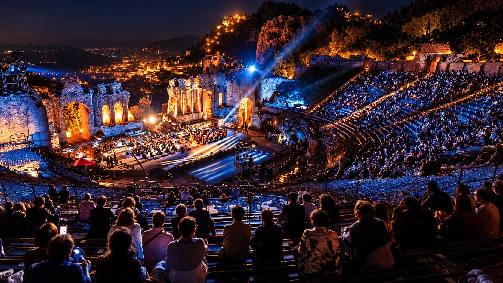 Teatro da concerto illuminata di blu con un grande palco, artisti e un pubblico seduto in primo piano.