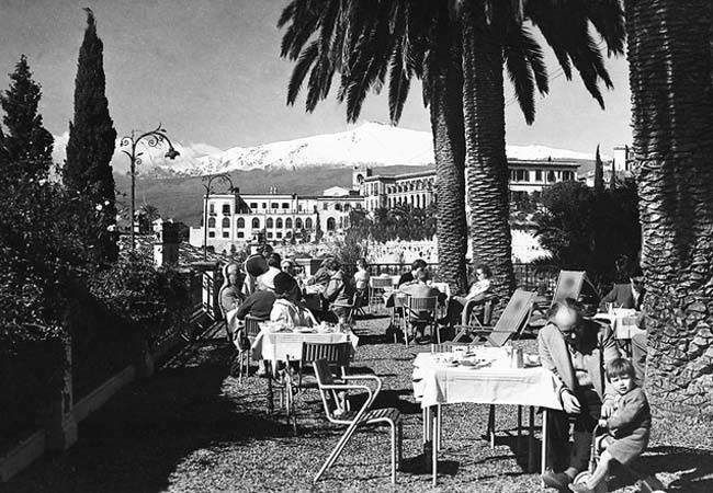 Terrasse de café avec palmiers, tables et personnes assises dans une cour ensoleillée.