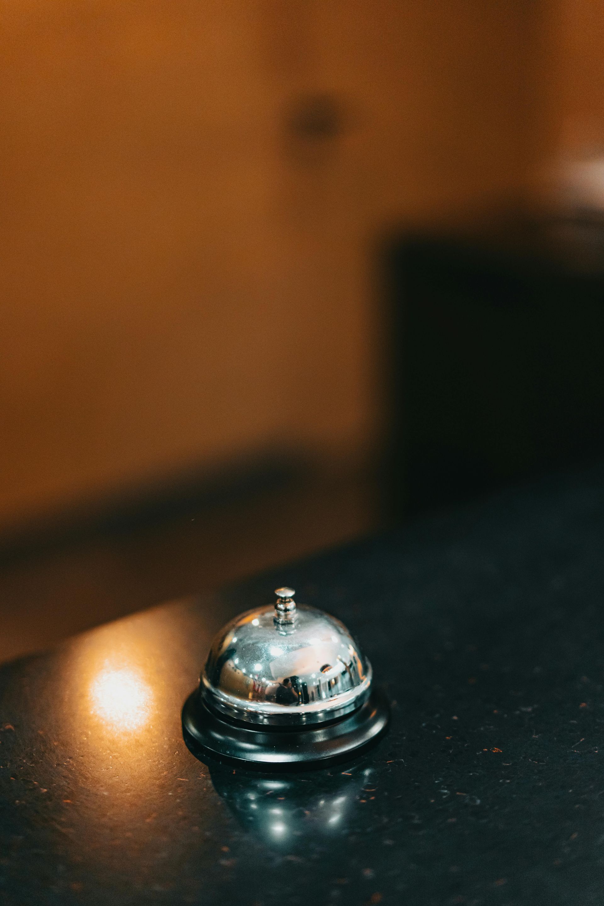 Piccola campanella di servizio in argento su un piano di lavoro scuro con uno sfondo sfocato dai toni caldi.