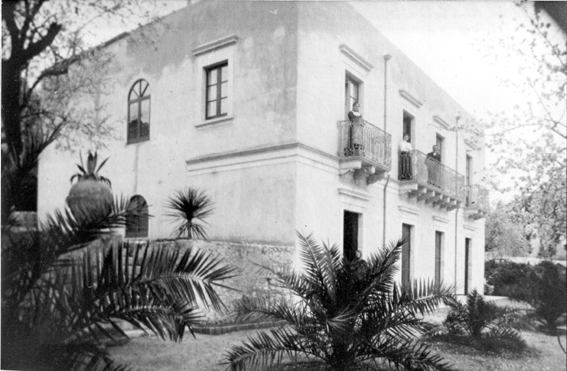 Black-and-white view of a two-story house with balconies and palm trees in the foreground