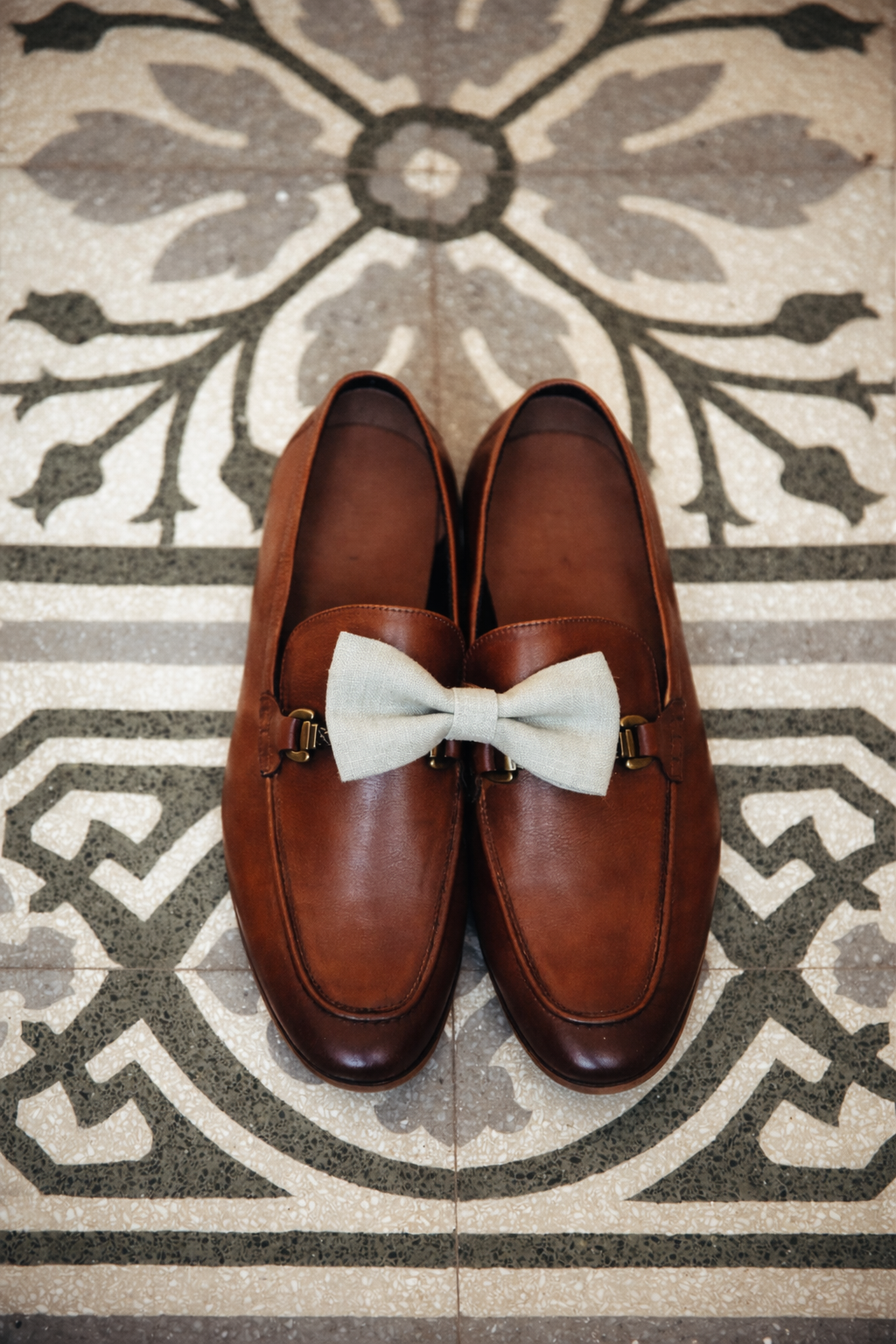 Brown leather loafers with a light bow tie, placed on a patterned rug.