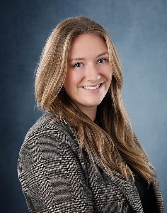Woman with long blonde hair smiles, wearing a plaid blazer, against a blue background.