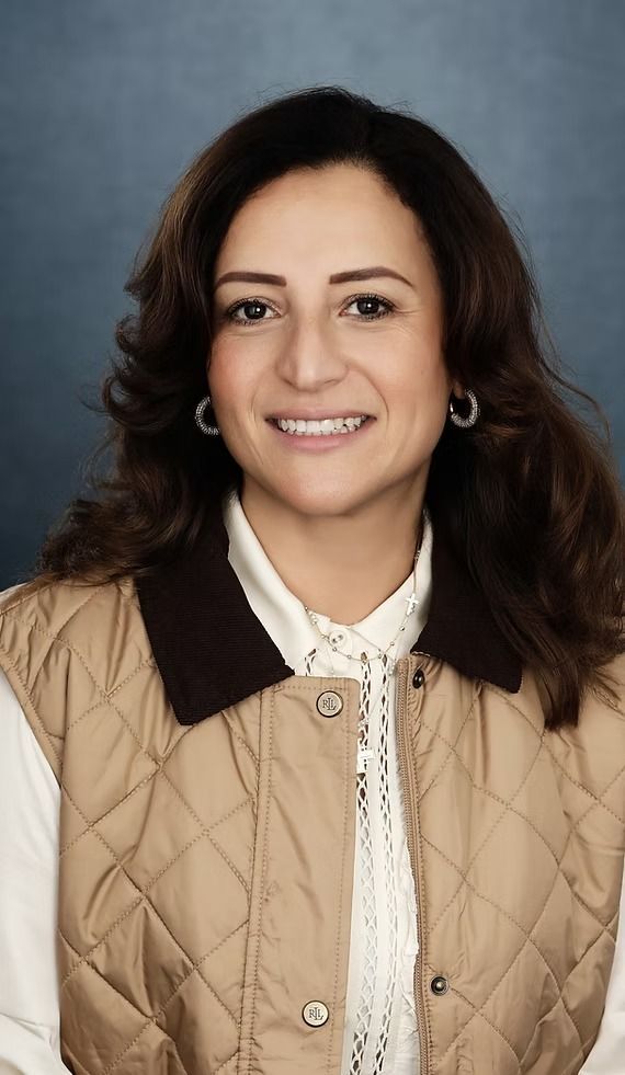 Woman in quilted tan vest and white shirt smiles, looking at camera against a blue background.