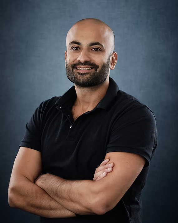 Man with a beard, arms crossed, smiling. Wearing black polo shirt, against a blue background.