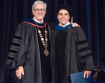 Two men in academic robes at a graduation ceremony. The man on the right holds a diploma, smiles, and has an arm around him.