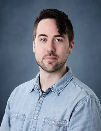Man with dark hair and a short beard, wearing a denim shirt, looking at the camera. Against a blue backdrop.