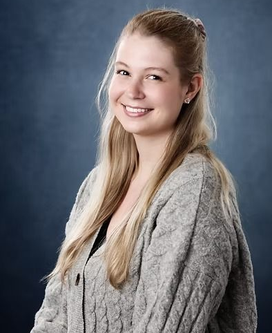 A smiling person with long blonde hair wearing a grey cable-knit sweater against a mottled dark blue background.