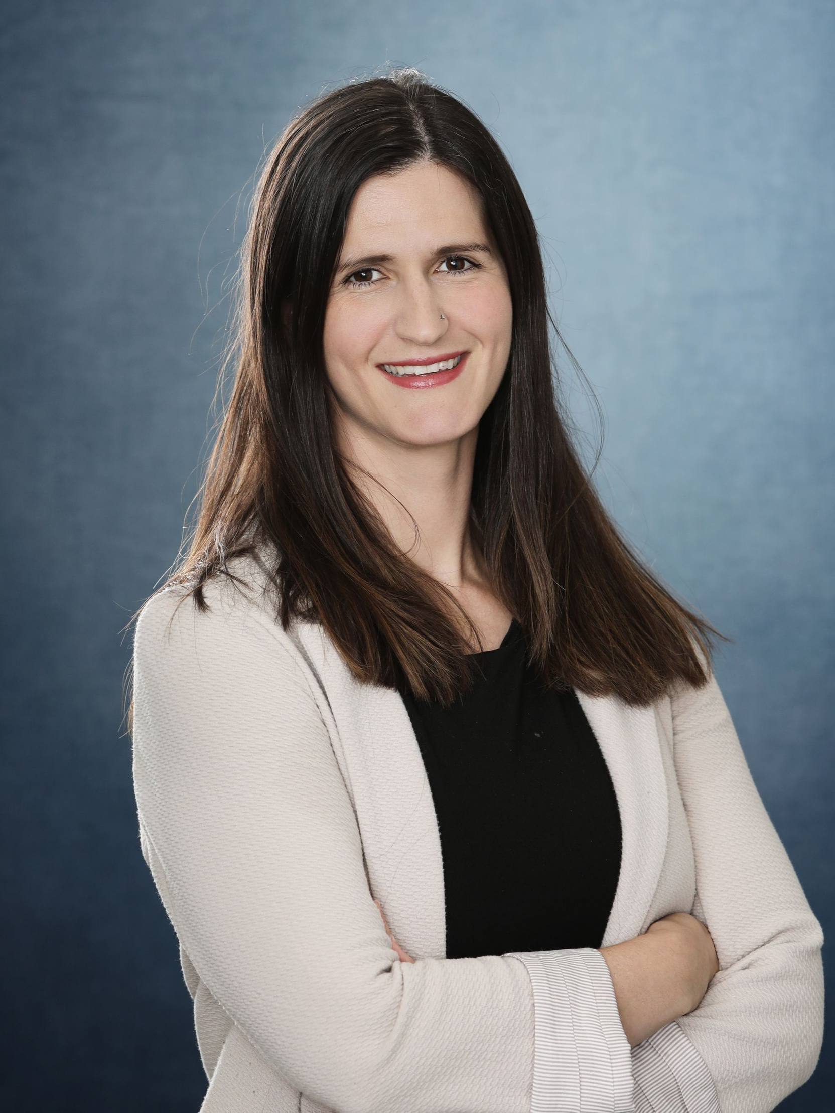Woman with long dark hair, arms crossed, wearing a tan blazer over a black top, against a blue background.