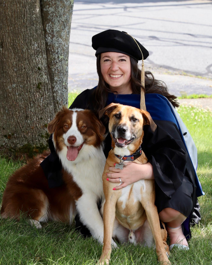Dr.Burns in graduation cap and gown sitting with two dogs on grass by a tree, all smiling.