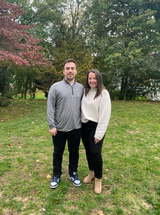 Dr. Burns and her partner, standing on grass in a park, smiling, with trees and autumn leaves behind them