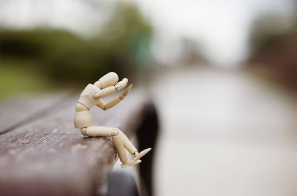 Wooden mannequin on a park bench, head in hands, slumped posture; blurred path in background.