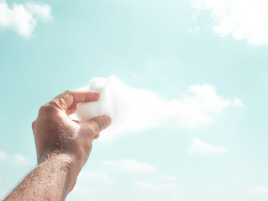 Hand holding a small, fluffy white object against a bright blue sky with scattered clouds.