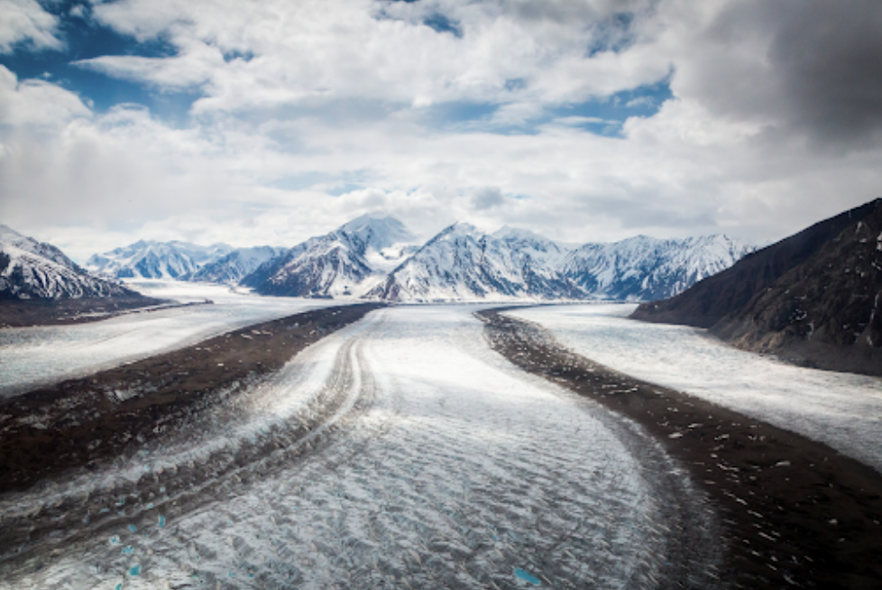 Glacier with dark rock-covered lateral moraines flowing between mountains under cloudy sky.