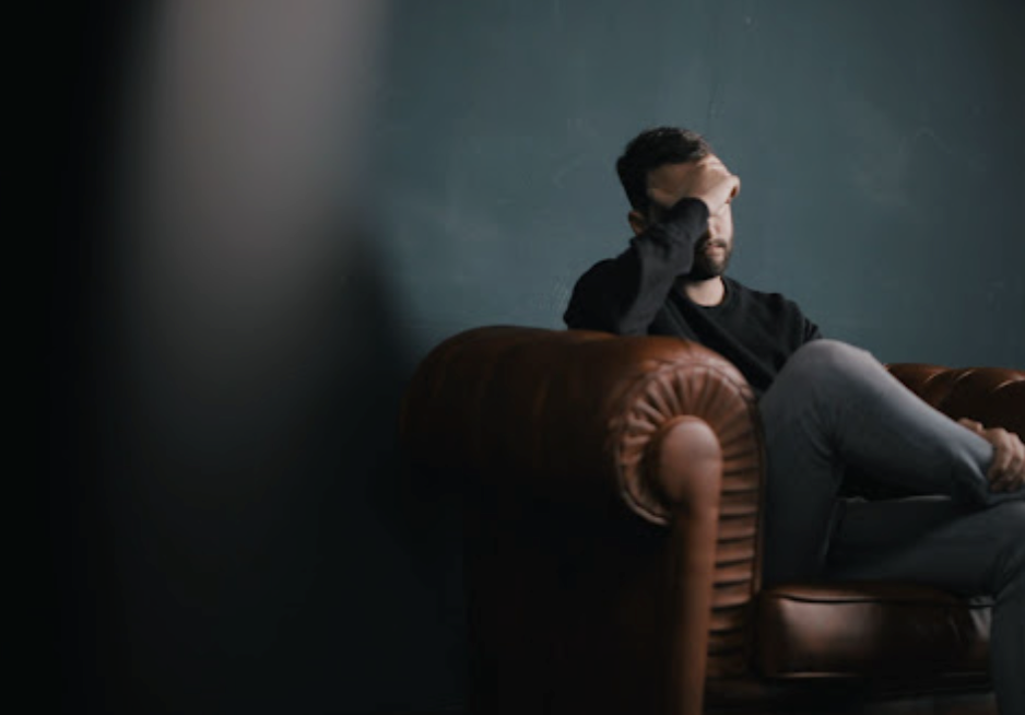 Man sitting in a leather armchair, face in hand, in a dimly lit room.