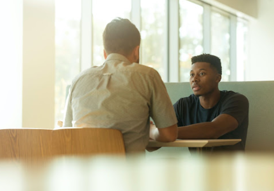 Two men sit at a table, conversing indoors. One man faces the camera, listening intently.