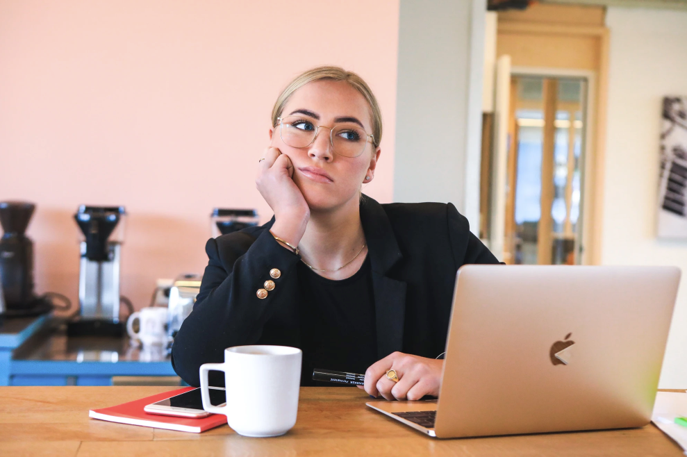 Woman in glasses, black blazer, and bored expression sits at a laptop with a coffee cup and notepad.