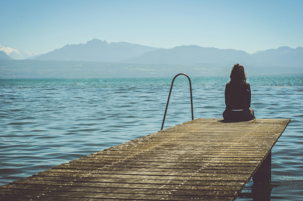 Woman seated at the end of a wooden dock, facing tranquil water and mountains on a sunny day.