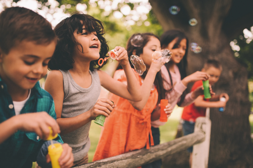 Children blowing bubbles outdoors. Bright colors, wooden fence, smiling faces.