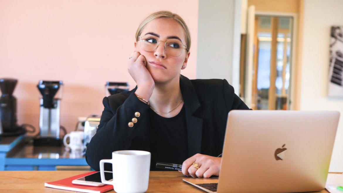 Woman with glasses looking bored, rests head on hand at desk with laptop, coffee mug.