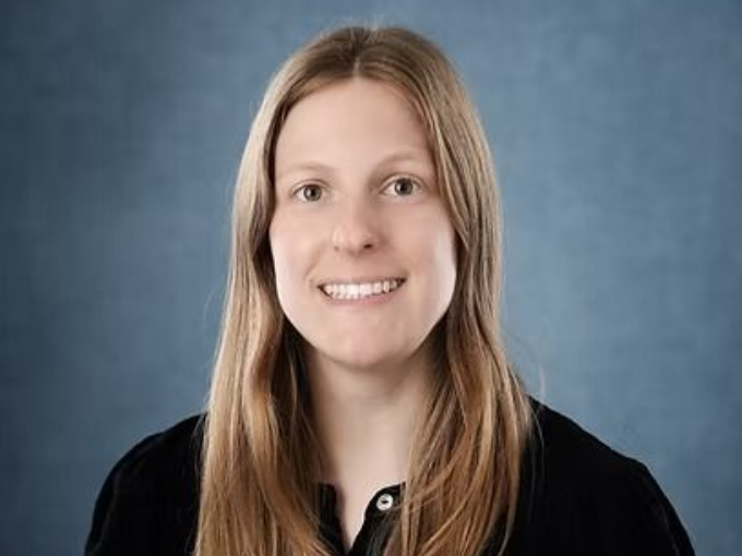 Woman with long blonde hair smiles, wearing a black shirt against a blue background.