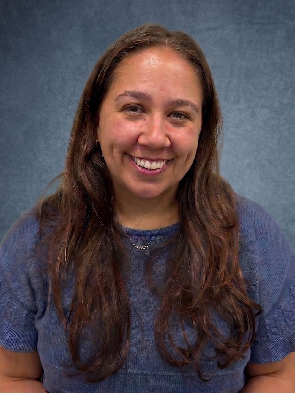 Smiling person with long brown hair in a blue shirt against a dark textured background