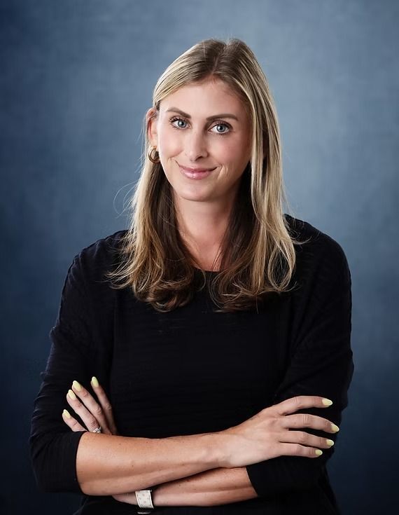 Woman with blonde hair wearing a black shirt, arms crossed, smiling, against a blue background.