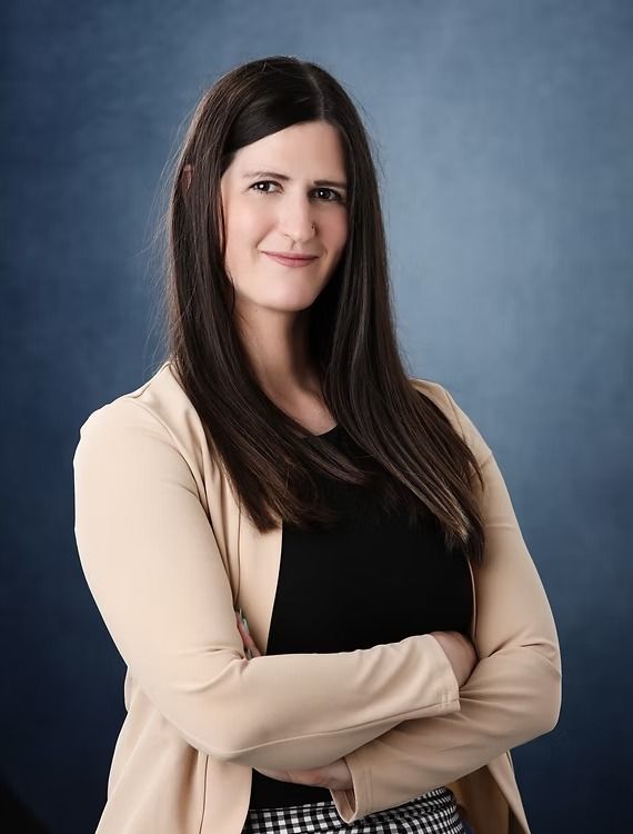 Woman in tan blazer and black top with arms crossed, smiling, blue background.
