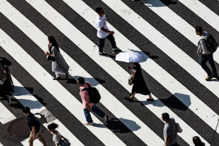 People crossing a diagonal zebra-striped street in daylight, one person uses an umbrella.