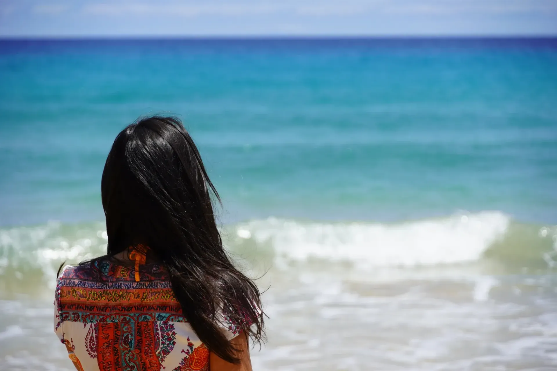Person with long dark hair looking at turquoise ocean.