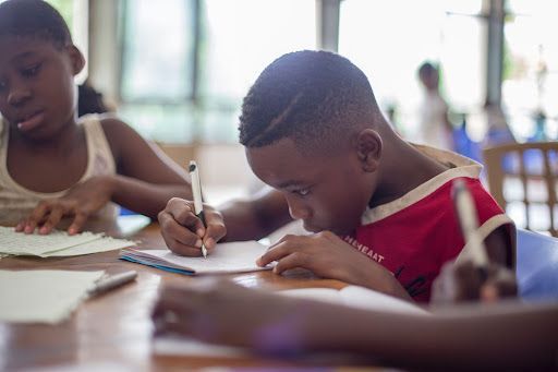 Children writing at a table; boy in red shirt focuses on paper.