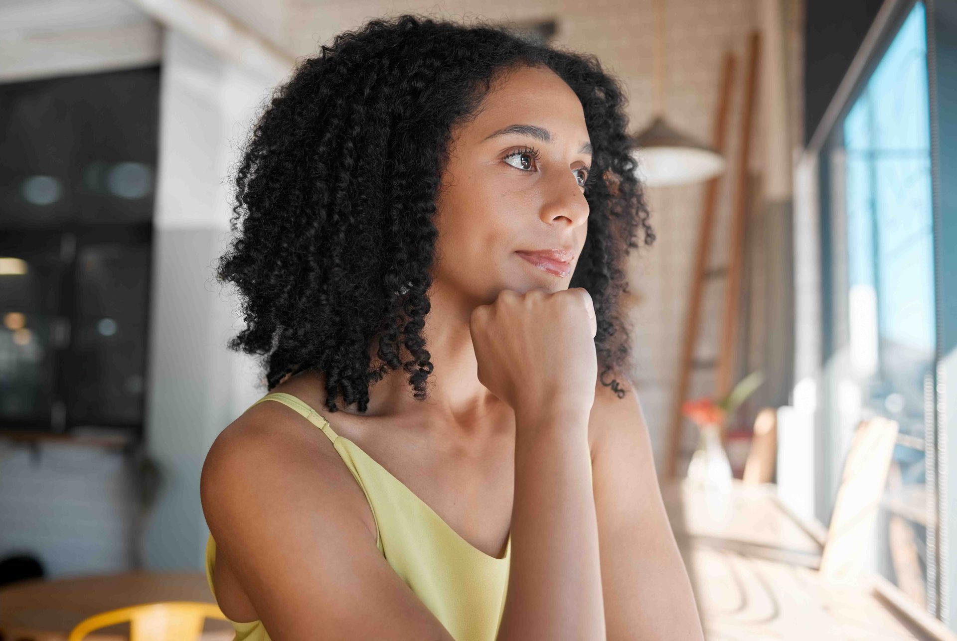 Woman with curly hair looking out a window, resting chin on hand, wearing a yellow top.