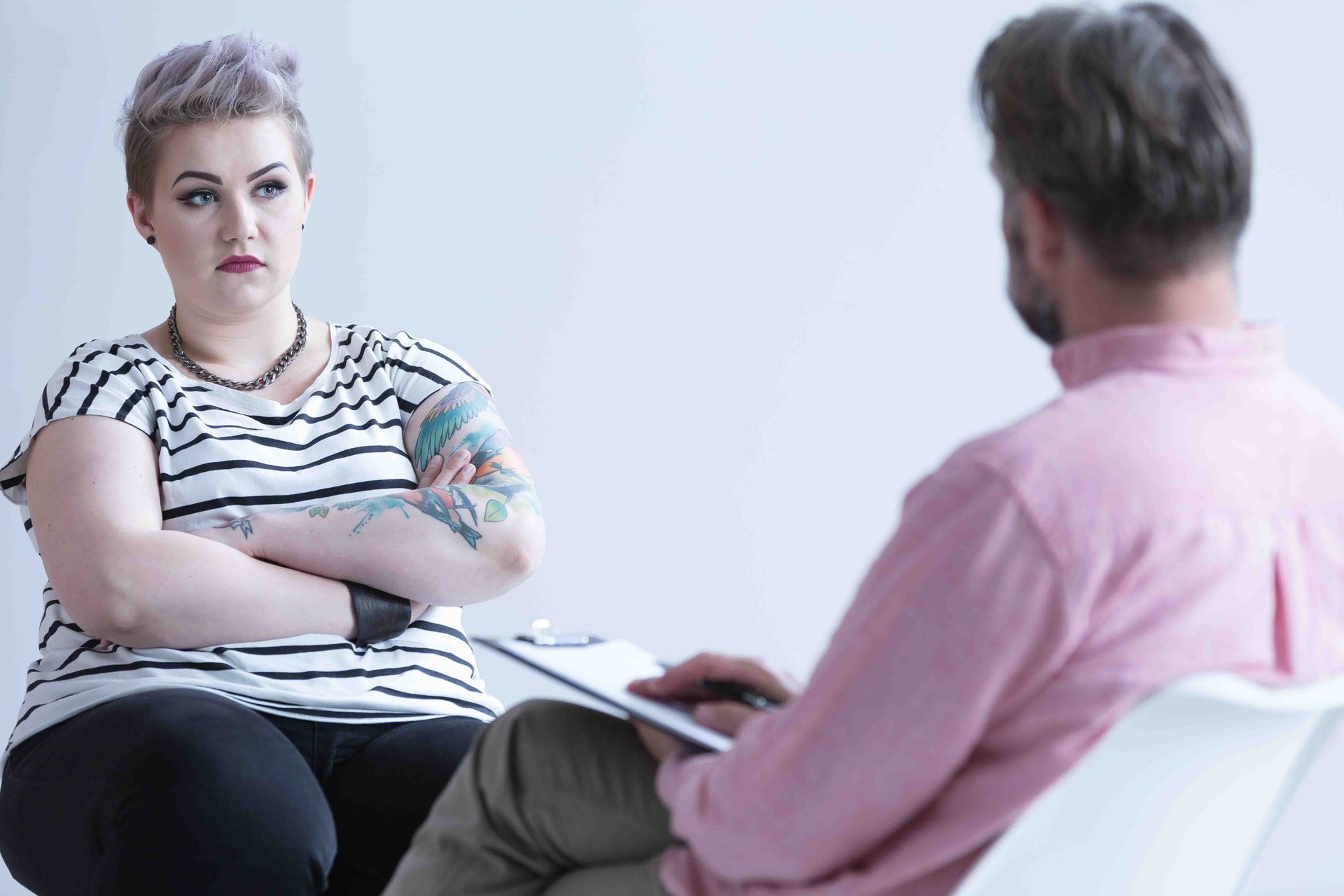Woman with crossed arms, looking displeased, talking to person holding a clipboard in a counseling setting.