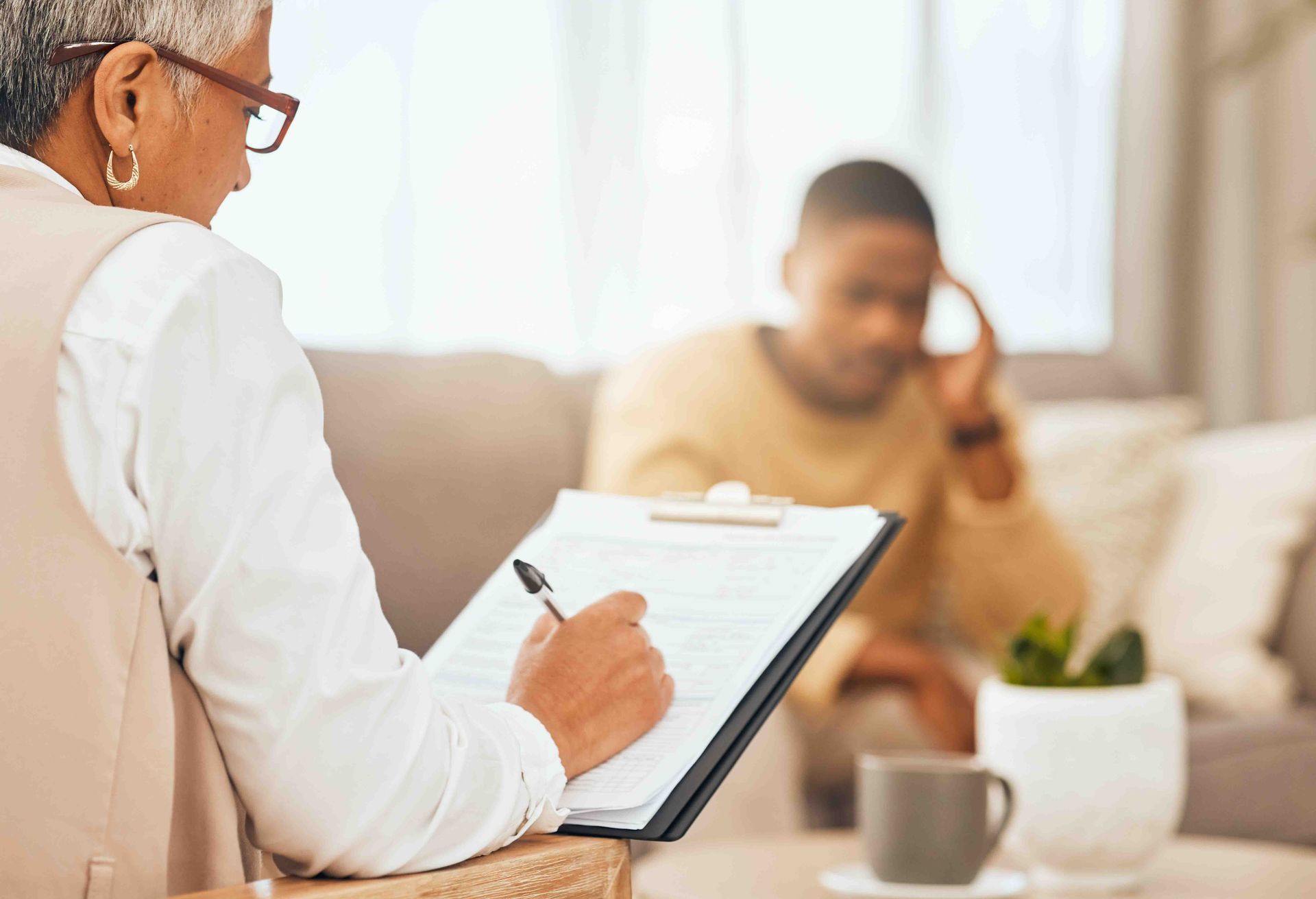 Therapist taking notes while client sits on a couch, holding their head. Indoor setting.