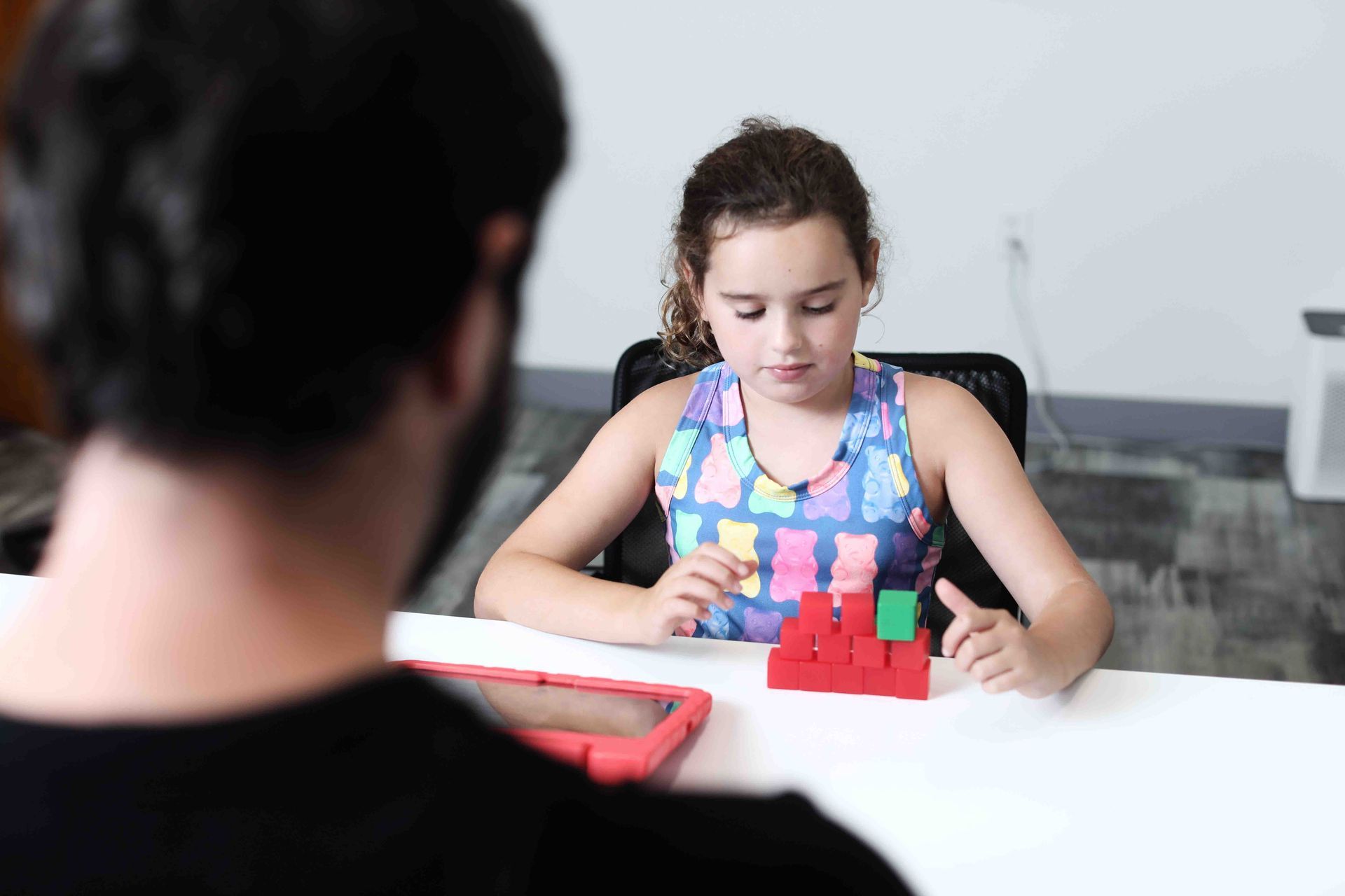 Girl building with blocks at a table, supervised by a person.
