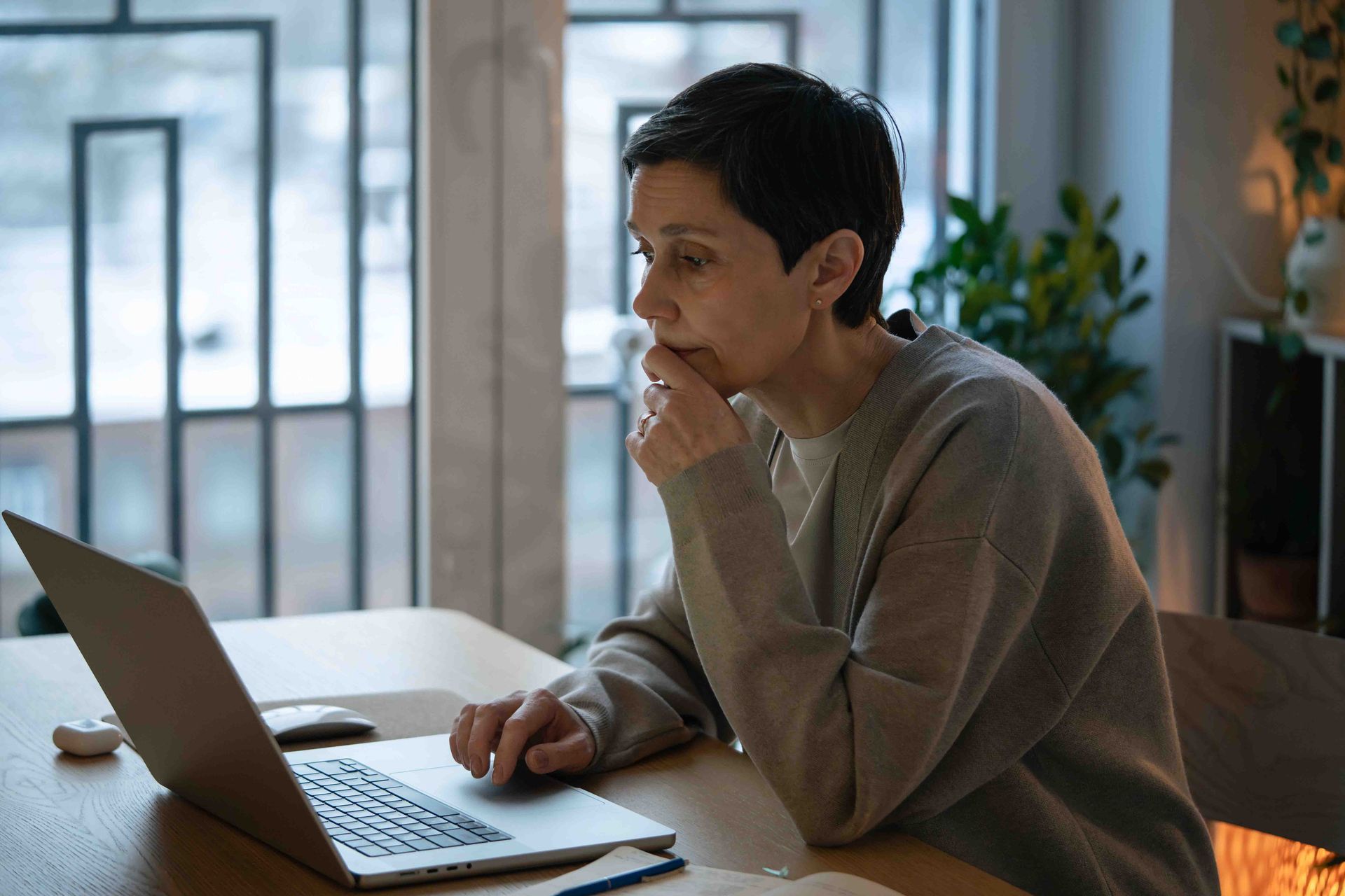 Woman using laptop at a table, thoughtful expression, hand on chin, indoors near a window.