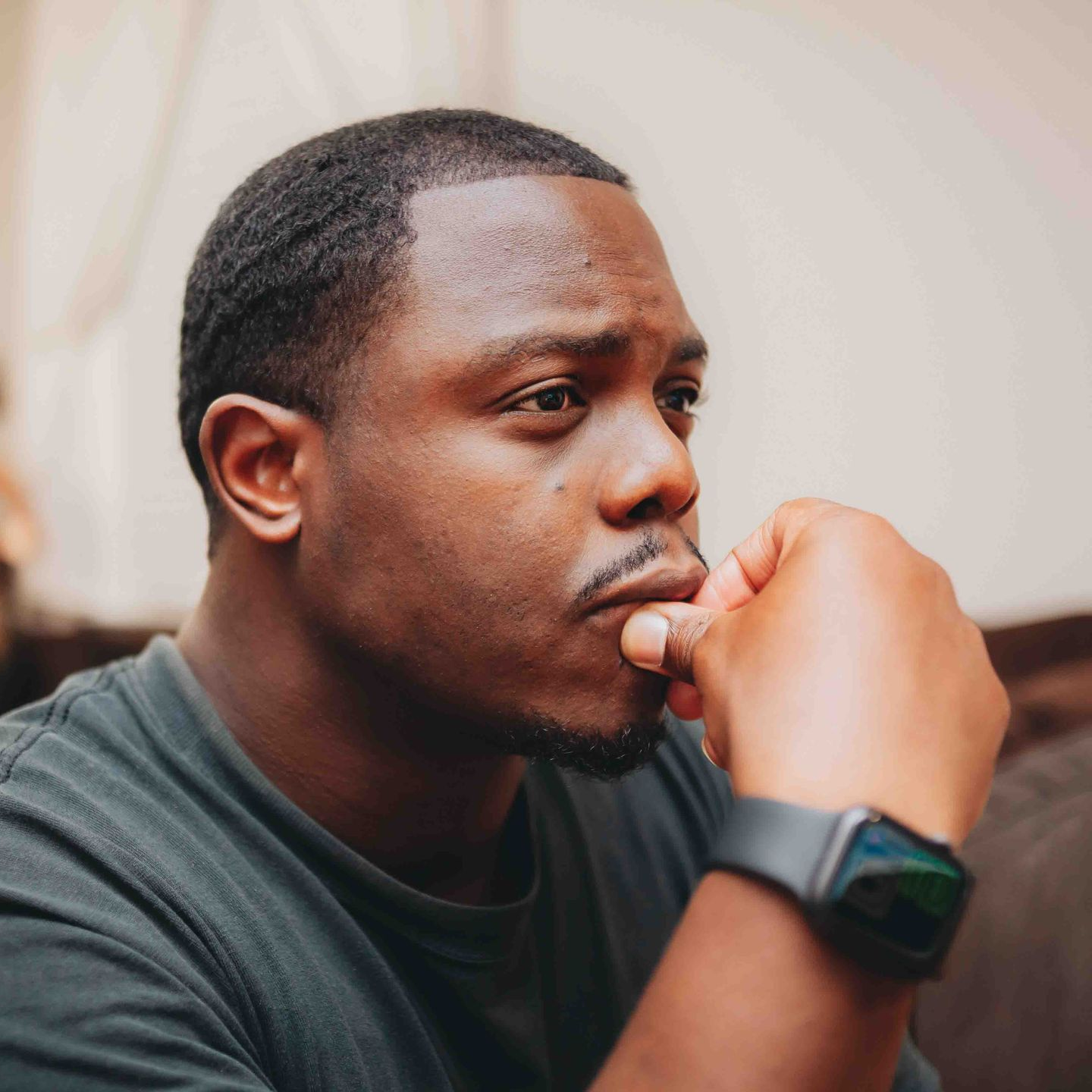 Man with dark skin, wearing a smartwatch, thoughtfully rests chin on hand.