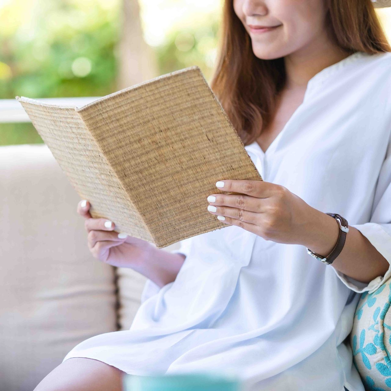 Woman reading a book with a tan cover while sitting outside. She's wearing a white shirt and a watch.