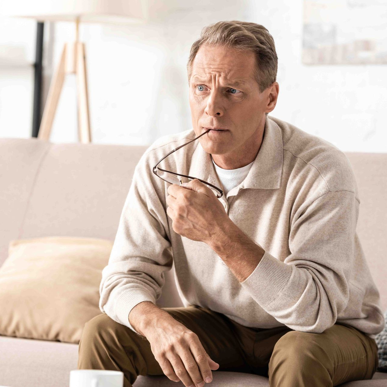 Man sitting on sofa, holding glasses, looking thoughtful. Living room setting.
