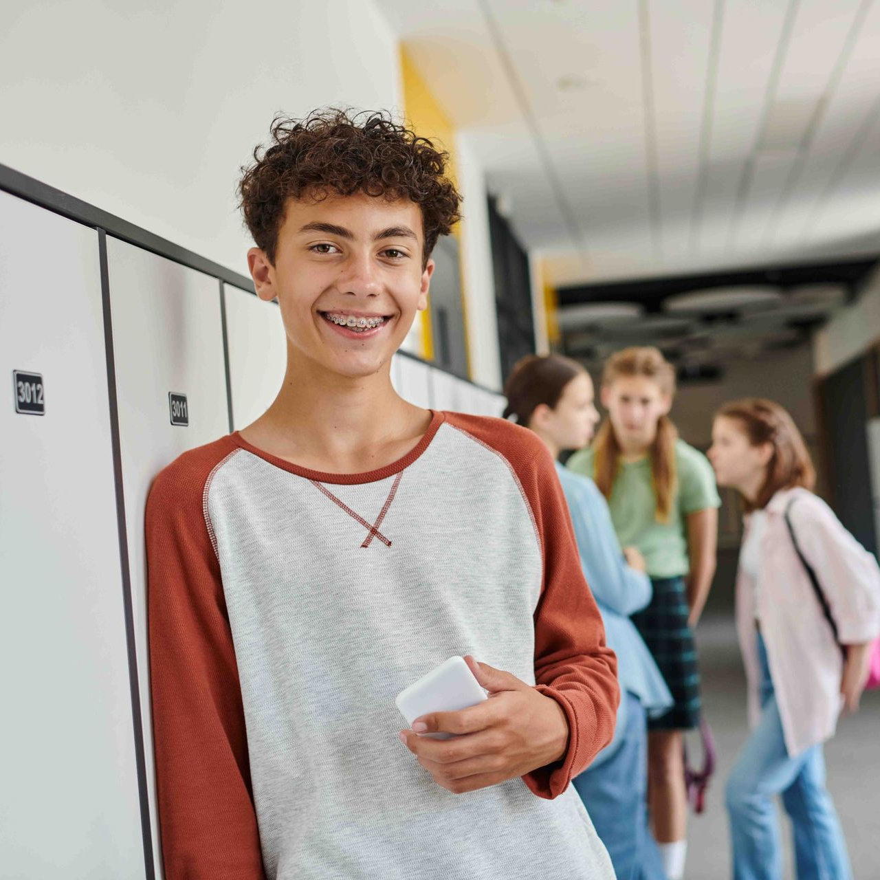 Teen with braces, leaning against lockers in school hallway, smiling, holding a phone.