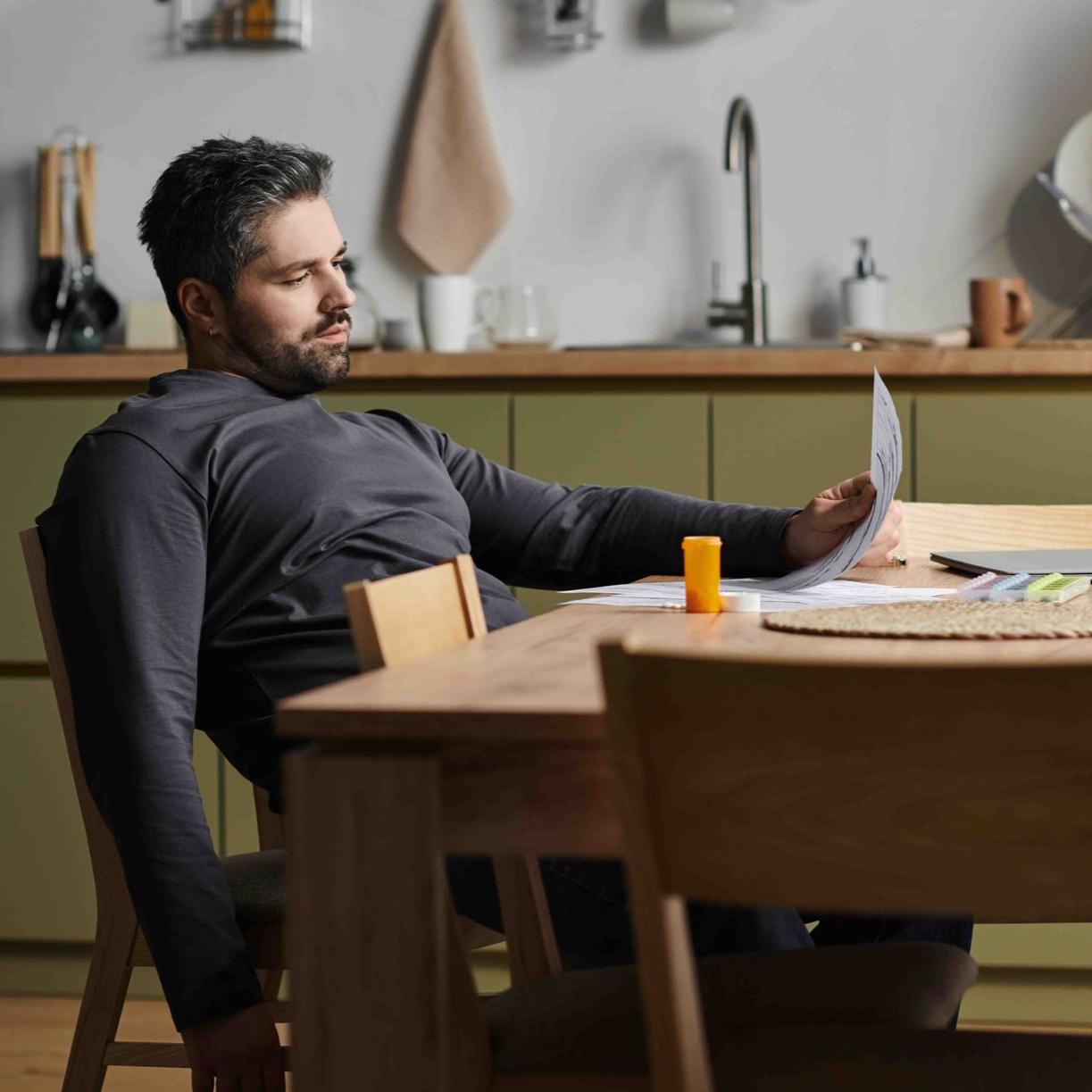 Man at a kitchen table, looking at papers. He holds a pen and sits near a small orange tube.