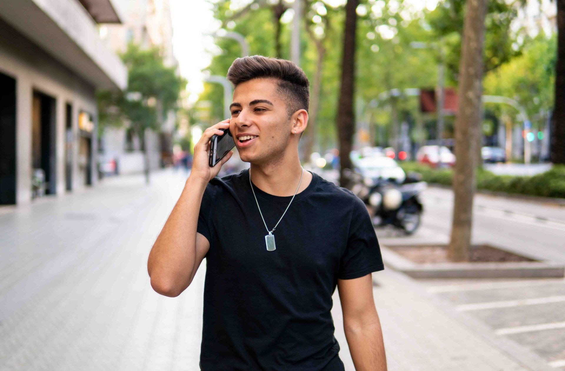 Person walking on sidewalk, talking on a cell phone. Green trees and buildings in the background.
