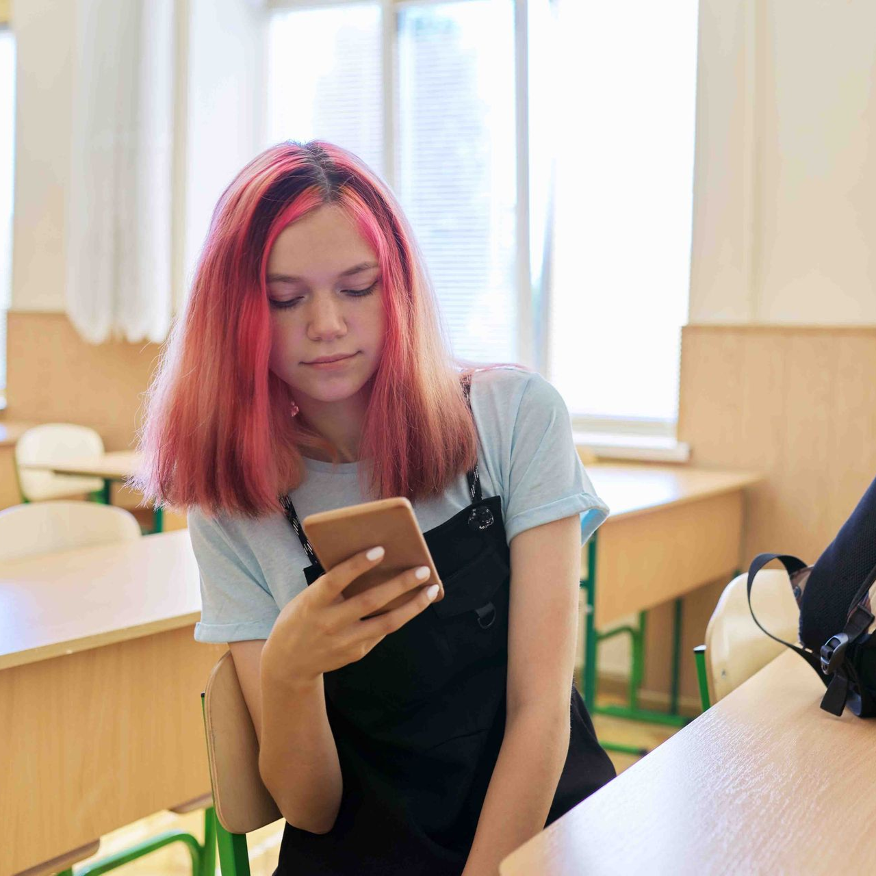 Teen with pink hair looks at phone in classroom.